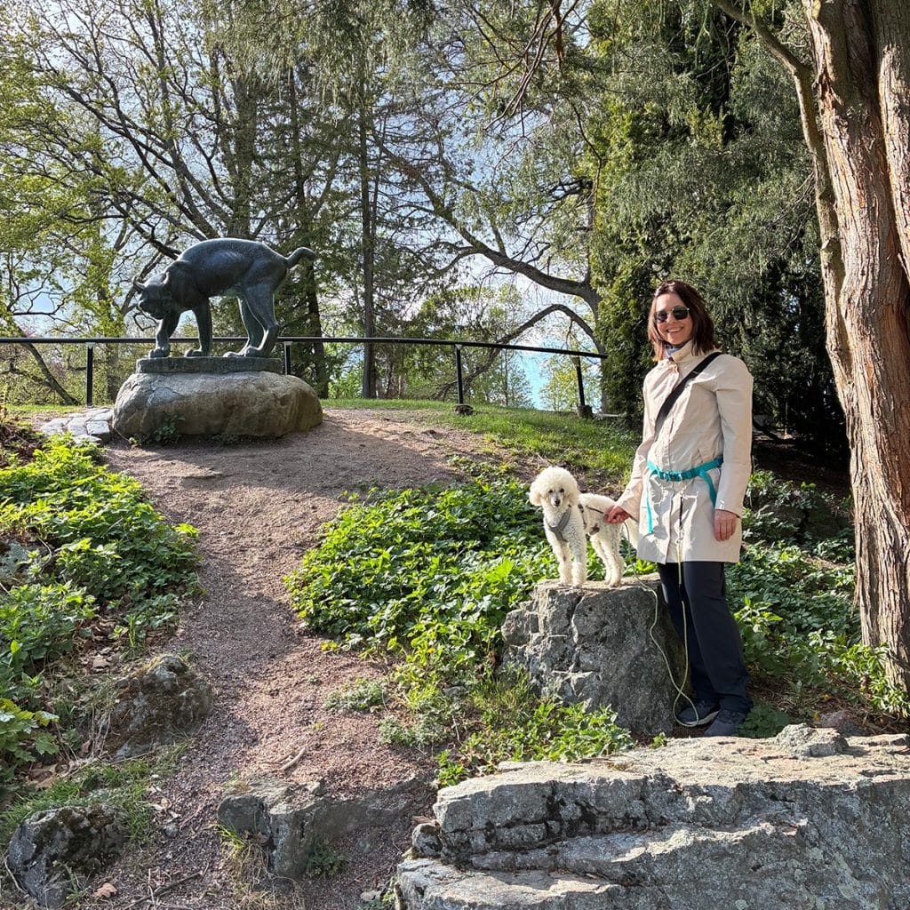 Kvinna med vit hund i en grönskande park framför skulptur av ett lodjur.