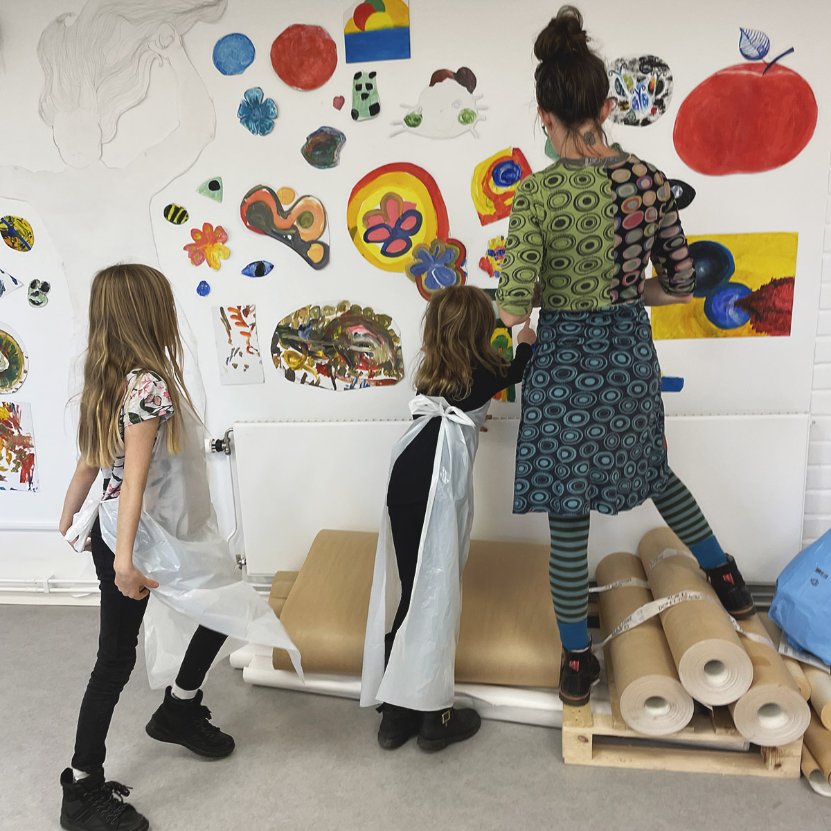 Två barn och en vuxen sätter upp målningar på en vägg.