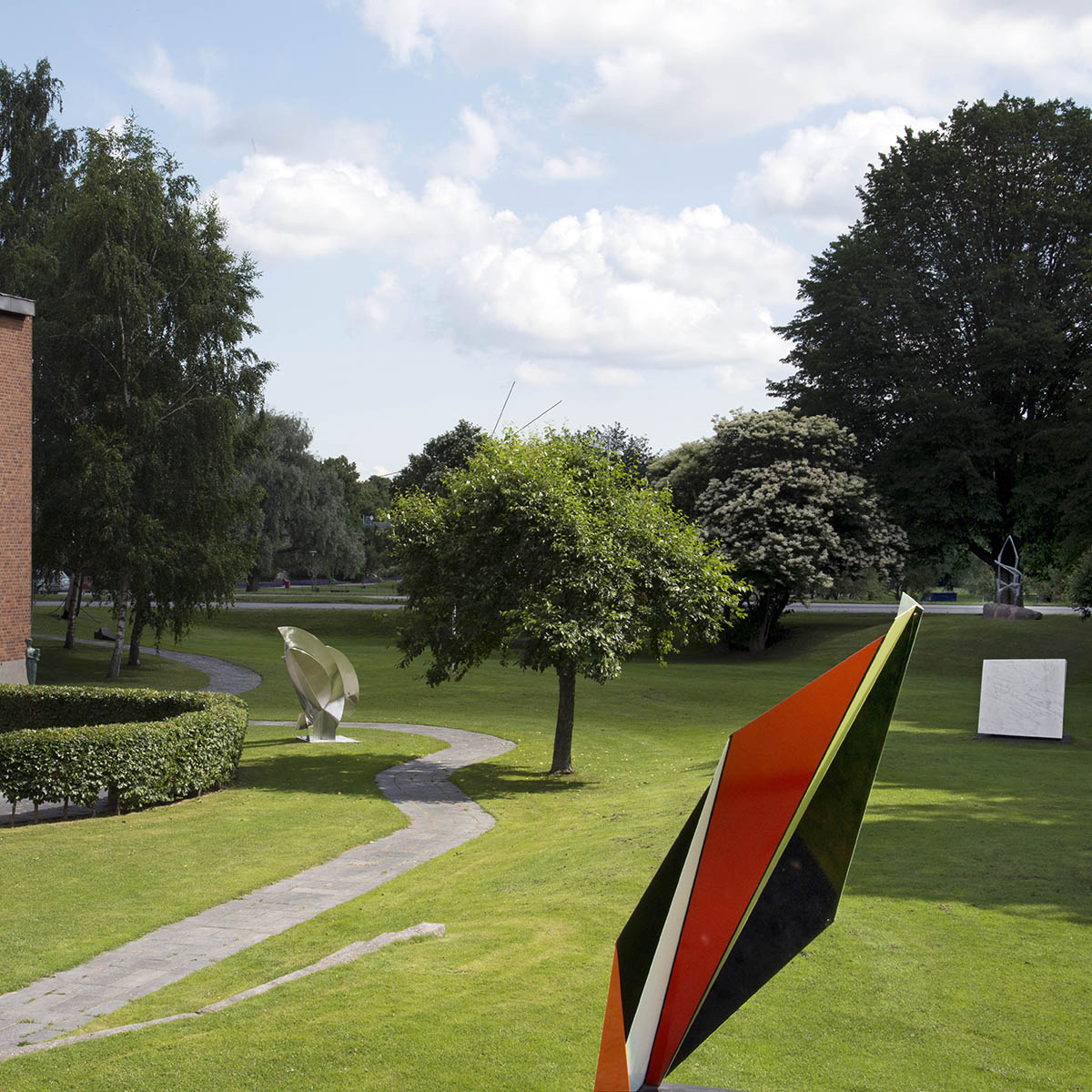 Foto på Konstmuseets Skulpturpark med Jacob Dahlgrens röda skulptur i förgrunden. I bakgrunden syns gräs, träd och fler skulpturer.