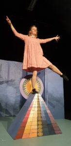 En tjej i rosa klänning balanserar på en pyramid med många färger, inspirerad av Hilma af Klints målningar. 